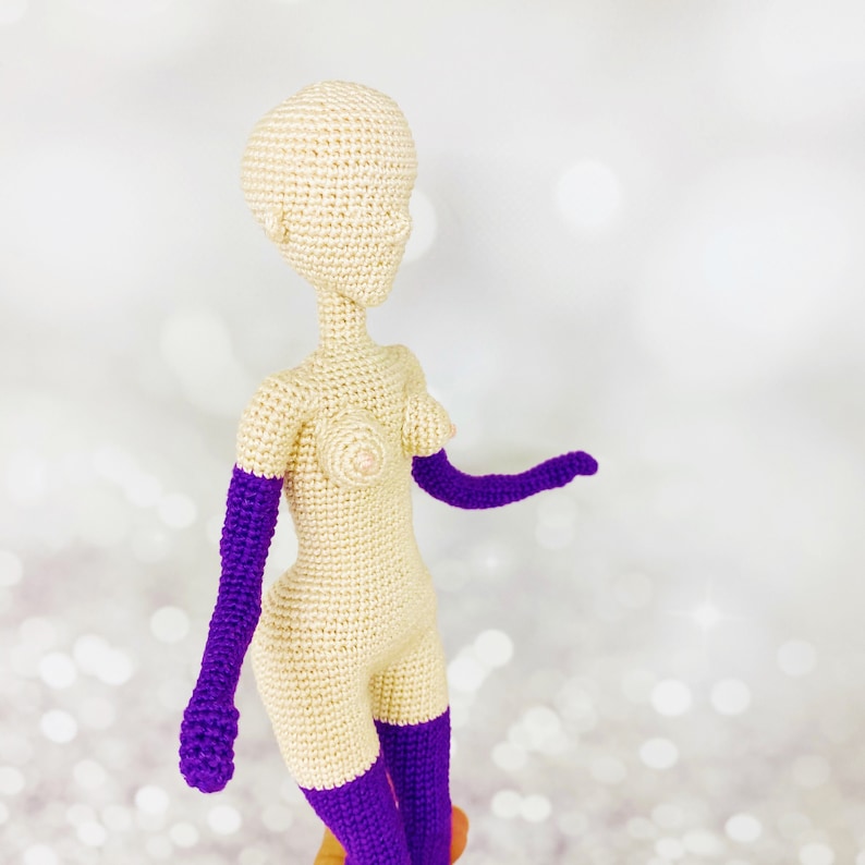 May include: A crocheted doll with a nude body and purple arms and legs. The doll is standing on a white background.