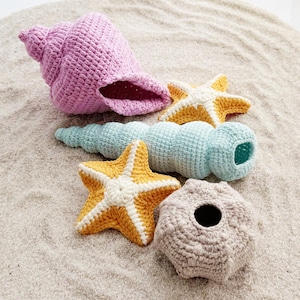 May include: A collection of five crocheted items including a large pink spiral seashell, a long light blue spiral shell, two yellow and white five-pointed starfish, and a textured beige sea urchin-shaped object with a central circular opening.