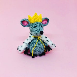 May include: A gray crocheted mouse wearing a white and black fur-trimmed robe and a yellow crown. The mouse is sitting on a pink background.