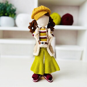 May include: A crocheted doll wearing a yellow hat, a striped sweater, a beige jacket, and green bell bottoms. The doll is holding a small camera and is standing on a white surface.