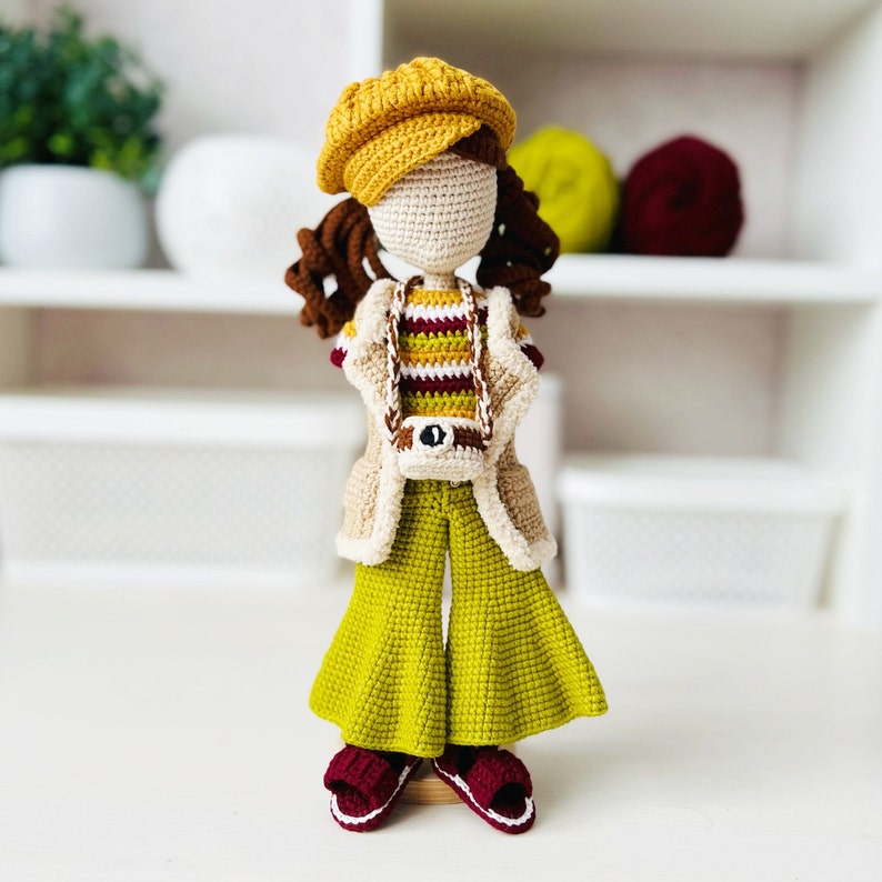 May include: A crocheted doll wearing a yellow hat, a striped sweater, a beige jacket, and green bell bottoms. The doll is holding a small camera and is standing on a white surface.