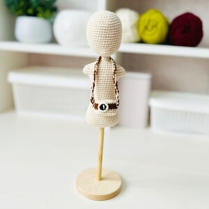 May include: A crocheted doll with a brown belt and a crocheted strap around its neck. The doll is standing on a wooden stand.