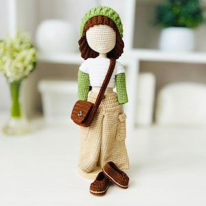 Può includere: Una bambola all'uncinetto con un cappello verde, una camicia bianca e pantaloni beige con una borsa marrone. La bambola è in piedi su una base di legno e ha scarpe marroni.