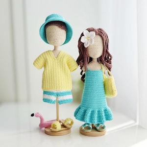 Op de afbeelding: Twee handgemaakte gehaakte poppen op houten voetstukken. De linkerpop draagt een blauwgroene vissershoed, een geel shirt en een gestreepte korte broek. De rechterpop heeft bruin haar met een witte bloem, een blauwgroene jurk en een gele tas.