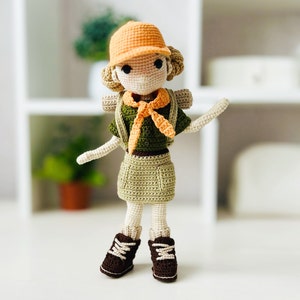May include: A crocheted doll wearing a green and orange scout uniform with a brown hat and brown boots. The doll has a crocheted backpack and a crocheted orange scarf.