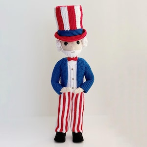 May include: A handmade Uncle Sam doll, crocheted with red, white, and blue yarn. The doll wears a tall striped hat, a blue jacket, and red and white striped pants. It has a white beard, a red bow tie, and black shoes. A patriotic decorative item.