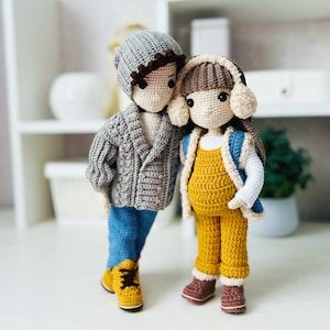 Peut inclure: Deux poupées en crochet portant des vêtements d'hiver. La poupée de gauche porte un pull gris, un jean bleu et un chapeau gris. La poupée de droite porte une combinaison jaune, un pull blanc et un chapeau marron avec des oreillettes.