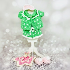 May include: A green and white crocheted baby doll outfit with a matching eye mask, a pink crocheted bunny, and a pair of pink crocheted slippers.