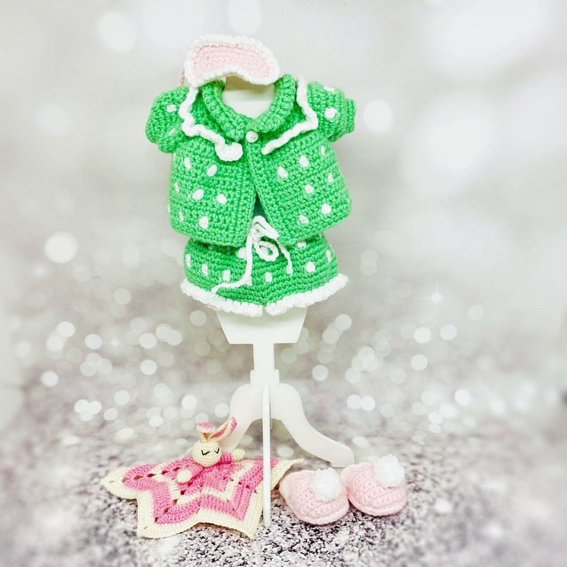 May include: A green and white crocheted baby doll outfit with a matching eye mask, a pink crocheted bunny, and a pair of pink crocheted slippers.