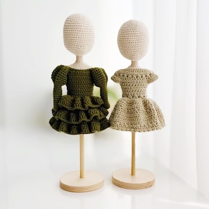 May include: Two crochet dolls with round, cream-coloured heads mounted on wooden stands. The left doll wears a dark olive green dress with long, puffed sleeves and a tiered, ruffled skirt. The right doll wears a light beige dress with short sleeves.