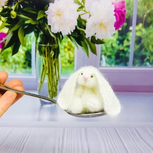 May include: A white crocheted bunny rabbit sits on a silver spoon. The bunny has pink ears and a pink nose.