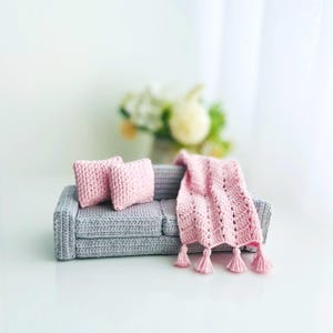 May include: A miniature gray crochet sofa with two pink crochet pillows and a pink crochet blanket with tassels. The sofa is a small-scale representation of a living room furniture set.