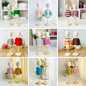 Op de afbeelding: Een verzameling miniatuur gehaakte truien in verschillende kleuren en patronen, tentoongesteld op houten mannequin vormen. De truien bevatten strepen, getextureerde breisels en bloemmotieven. De mannequins staan op houten standaards.