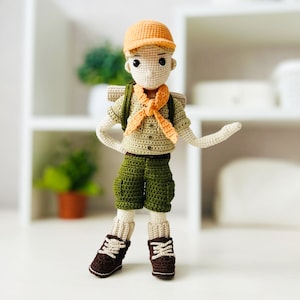 May include: A crocheted doll wearing a green and brown scout uniform with an orange neckerchief. The doll is wearing a brown hat and has a backpack.