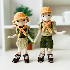 Op de afbeelding: Twee gehaakte poppen gekleed in scout-uniformen. De poppen dragen oranje hoeden, groene shorts en oranje dasjes. De poppen houden elkaars hand vast en glimlachen.