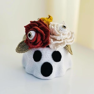 Puede incluir: Un fantasma de ganchillo blanco hecho a mano con detalles negros de ojos y boca. El fantasma está adornado con flores de ganchillo rojas, blancas y amarillas, con hojas verdes. Una decoración de Halloween única y caprichosa.