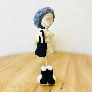 May include: Crocheted doll wearing a blue beret, black boots, and a black tote bag. The doll is standing on a wooden base.
