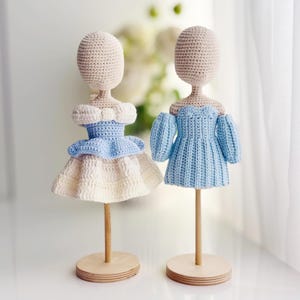 May include: Two crochet dolls on wooden stands. The left doll wears a light blue and white dress with a peplum waist. The right doll wears a solid light blue dress with off-the-shoulder puff sleeves. Both dolls have beige yarn heads and bodies.