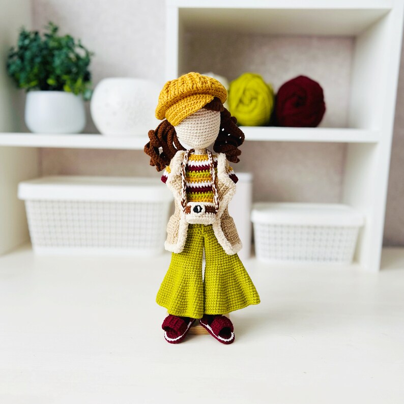 May include: A crocheted doll with brown hair, wearing a yellow and brown striped shirt, a yellow jacket, and green bell bottom pants. The doll is wearing a yellow hat and brown shoes.