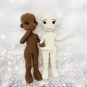 May include: Two crocheted dolls, one brown and one white, standing side by side. Both dolls have black eyes and red lips. The brown doll is holding its hand to its chin. The white doll has one arm outstretched.