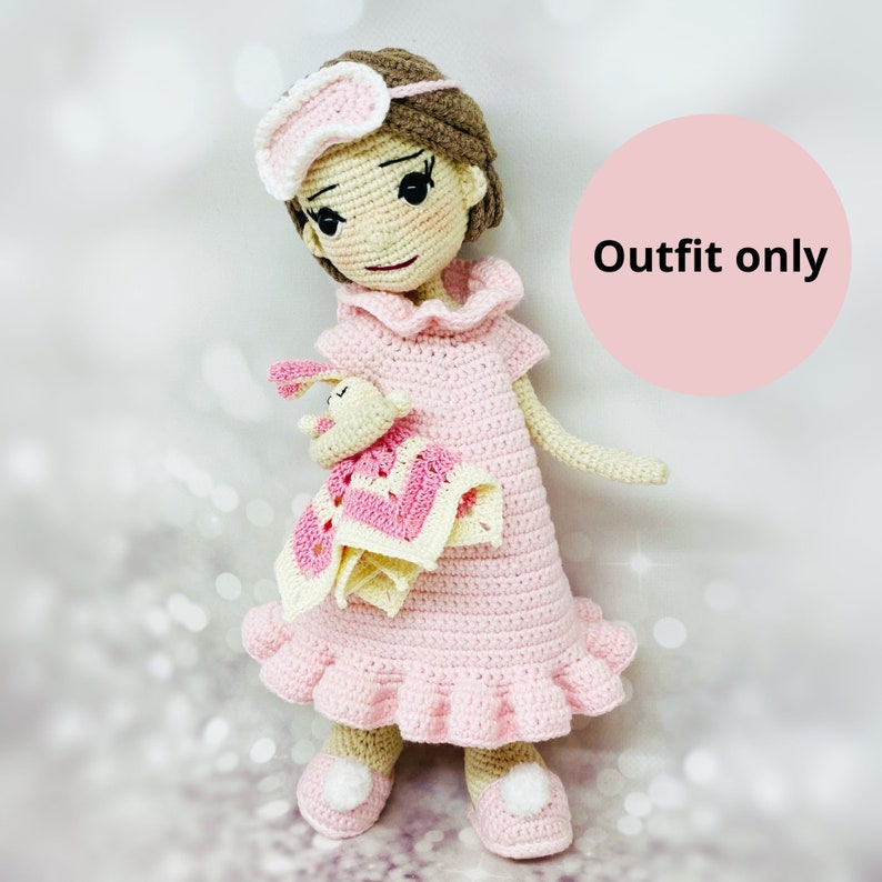 May include: A pink crocheted doll wearing a pink nightgown and a white eye mask. The doll is holding a pink and yellow crocheted blanket. The doll is standing on a white surface. Outfit only.