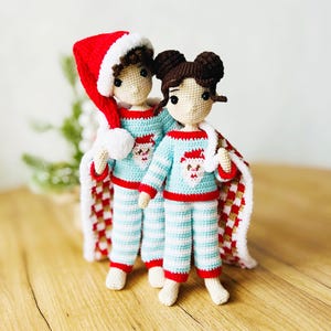 May include: Two crocheted dolls wearing blue and white striped pyjamas with red trim and Santa Claus designs. The dolls have brown hair and are holding a plaid blanket.