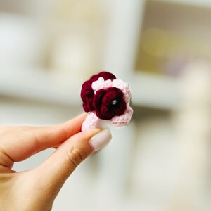 May include: A small crocheted bouquet of pink and burgundy flowers with a pearl accent.