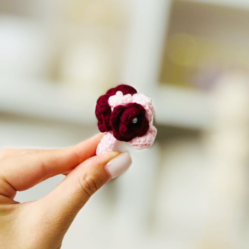 May include: A small crocheted bouquet of pink and burgundy flowers with a pearl accent.