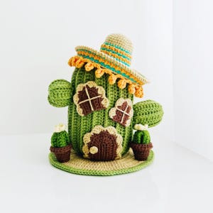 May include: Handmade crochet cactus house with a sombrero. The green cactus has brown windows and a door, with two small cactus plants in pots. The sombrero is tan with orange and turquoise trim. The cactus house sits on a tan and green base.