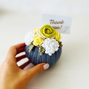 May include: A crocheted flower arrangement in a gray pot. The flowers are yellow, green, and white. The pot is decorated with a crocheted design. The arrangement is sitting on a white surface. The text "Thank you!" is visible in the background.