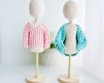 Könnte beinhalten: Zwei gehäkelte Puppenkörper auf Holzständern, jeder mit einem anderen gehäkelten Pullover. Ein Pullover ist rosa und der andere türkis.