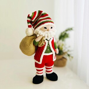 May include: A crocheted Santa Claus doll with a white beard and a red and green striped hat. He wears a red suit with white trim, black boots, and carries a tan sack. The doll is a festive Christmas decoration.