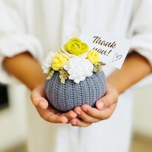 May include: A gray crocheted sphere with yellow, white, and green crocheted flowers on top. A small card with the text "Thank you!" is attached to the flowers.