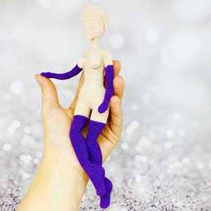May include: A crocheted doll with a nude body and purple limbs. The doll is posed sitting with its legs crossed.