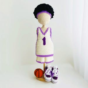 May include: A handcrafted crochet doll wearing a basketball uniform. The doll has a white jersey and shorts with purple trim and the number 1. It has a black afro, purple headband, and purple and white trainers. An orange basketball is also present.