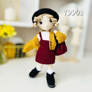 Può includere: Una bambola all'uncinetto con un cappello nero, un cardigan giallo, un vestito rosso e scarpe nere. La bambola ha i capelli biondi e gli occhi marroni. La bambola è in piedi su una superficie bianca. 1990s