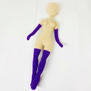 May include: A crocheted doll with a nude body and purple arms and legs. The doll is lying on a white surface.