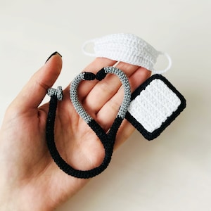 Peut inclure: Un stéthoscope, un masque facial et un petit objet carré en crochet dans une main. Le stéthoscope est noir et gris, le masque facial est blanc et l'objet carré est blanc avec une bordure noire.