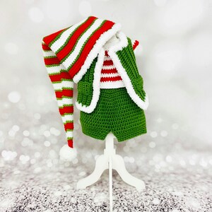Peut inclure: Un costume d'elfe crocheté vert et blanc pour bébé. La tenue comprend un chapeau, un gilet et un short. Le chapeau a une longue pointe pointue et des rayures rouges et blanches.