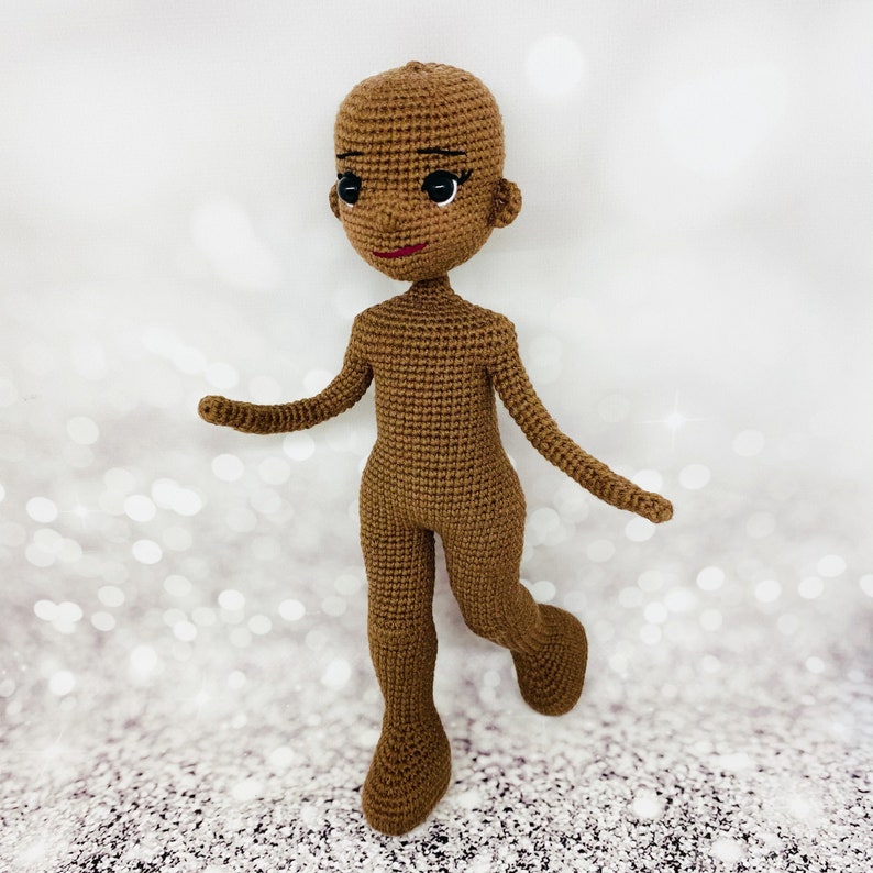 May include: A crocheted doll with brown skin, black eyes, and red lips. The doll is wearing a brown outfit and has a simple, minimalist design.