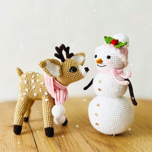 May include: A crocheted reindeer with brown antlers and a pink scarf stands next to a crocheted snowman wearing a pink hat with green holly. Both are on a wooden surface.