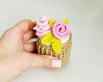 May include: A crocheted pincushion shaped like a cupcake with pink and green flowers. The pincushion is filled with white beads and has a brown crocheted base.
