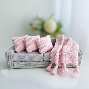 May include: Miniature gray crochet sofa with three pink crochet pillows and a pink crochet blanket with tassels. The sofa is a small-scale model, perfect for a dollhouse or craft project.