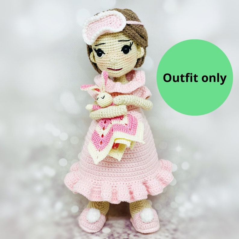 May include: A crocheted doll wearing a pink nightgown and a white eye mask, holding a crocheted bunny. The doll is standing on a white surface. "Outfit only" is written on a green circle in the background.
