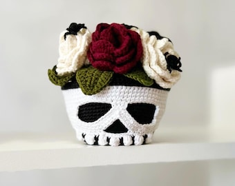 May include: A crocheted skull planter with a white and black skull design. The planter is decorated with white and burgundy flowers and green leaves.