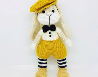May include: A crocheted bunny toy wearing a yellow beret, a white shirt with a black bow tie, yellow pants, and black and white striped socks. The bunny has a white face with black eyes and a black nose.