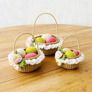 May include: Three handmade crochet baskets with handles, each filled with colorful crochet eggs and floral accents. The baskets are brown with white lace trim and are arranged on a wooden surface.