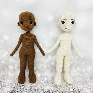 May include: Two crocheted dolls, one with brown skin and one with white skin. Both dolls have black eyes and red lips. The dolls are standing on a white background.
