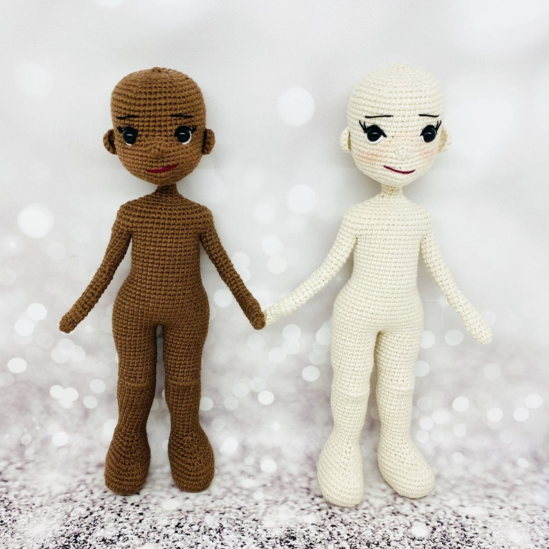 May include: Two crocheted dolls, one with brown skin and one with white skin. Both dolls have black eyes and red lips. The dolls are standing on a white background.