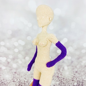 May include: A crocheted doll with a nude body and purple arms. The doll is made of yarn and has a simple design.